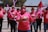 A group of people wearing pink shirts and masks participate in a demonstration or event. They are holding pink balloons and giving thumbs up gestures. A red flag is visible in the background along with trees and part of a building. The shirts have a logo or graphic on them.