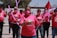A group of people wearing pink shirts and masks participate in a demonstration or event. They are holding pink balloons and giving thumbs up gestures. A red flag is visible in the background along with trees and part of a building. The shirts have a logo or graphic on them.