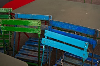 Several colorful, weathered folding chairs are arranged in a casual, overlapping manner. The chairs feature bold paint colors, including green and blue, with visible signs of wear and rust on the metal frames. In the background are simple tables, one appearing red and the other grey.
