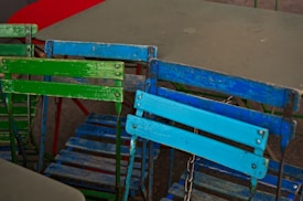 Several colorful, weathered folding chairs are arranged in a casual, overlapping manner. The chairs feature bold paint colors, including green and blue, with visible signs of wear and rust on the metal frames. In the background are simple tables, one appearing red and the other grey.