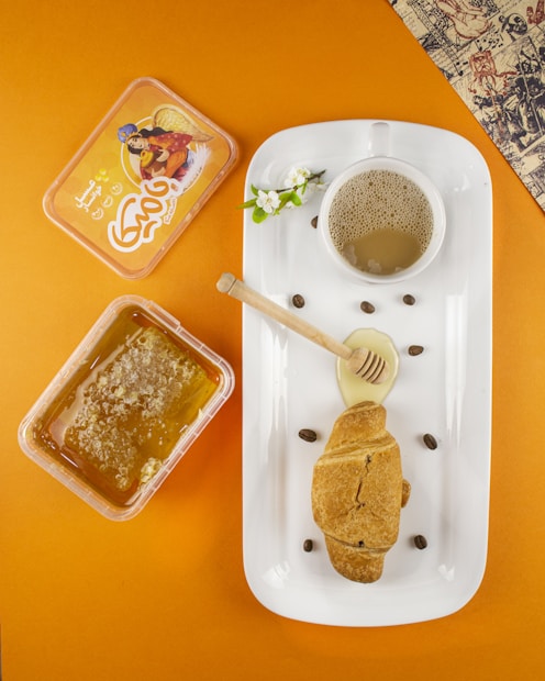 A white rectangular dish holds a cup of foamy coffee, a croissant, a honey dipper with honey, and some coffee beans scattered around. A small white flower adds a decorative touch. To the left, there is a plastic container of honeycomb. The background is a warm orange, and a corner of decorative paper is visible at the top right.