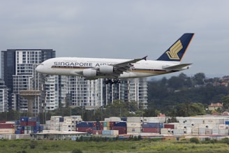A large airplane from Singapore Airlines is captured in mid-air, flying over an urban area. High-rise buildings and a landscape of trees are visible in the background, while shipping containers are stacked in the foreground.