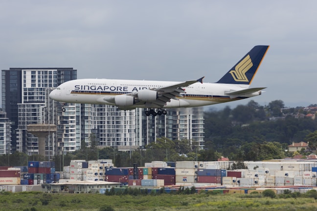 A large airplane from Singapore Airlines is captured in mid-air, flying over an urban area. High-rise buildings and a landscape of trees are visible in the background, while shipping containers are stacked in the foreground.
