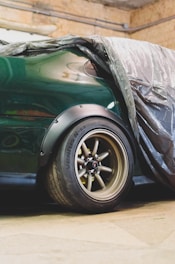 A partially covered green car is parked in a garage. The focus is on the rear section, showcasing the wheel with a visible tire marked with 'Ventus R-s3'. The car is draped with a plastic sheet, revealing the glossy surface.