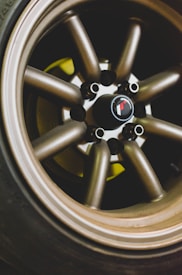 Close-up view of a multi-spoke alloy wheel with a metallic finish and a brand emblem at the center. The tire surrounding the wheel is visible, showing some wear. The lighting highlights the shiny texture of the metal and slight shading creates a sense of depth.