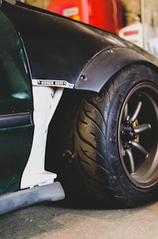 A close-up of a car's rear section featuring a large black tire with a distinctive tread pattern. The car has a dark green body with a flared fender and a decal that says 'Track Life.' The background is blurred, showing hints of red and beige colors.