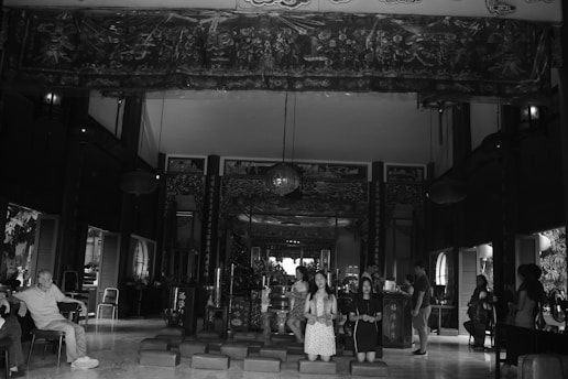 The interior of a traditional building with ornate decorations and hanging lanterns. Several people are inside, including two sitting on the left and a group of individuals kneeling in the center. The architecture features intricate woodwork and detailed patterns on the walls.