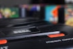 A close-up view of a Sega Mega Drive console, featuring the brand logo prominently. The black plastic exterior is in focus, with vibrant, blurry colors in the background suggesting vintage video game cartridges.