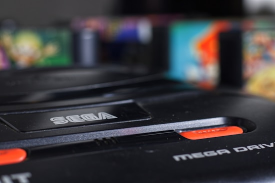 A close-up view of a Sega Mega Drive console, featuring the brand logo prominently. The black plastic exterior is in focus, with vibrant, blurry colors in the background suggesting vintage video game cartridges.
