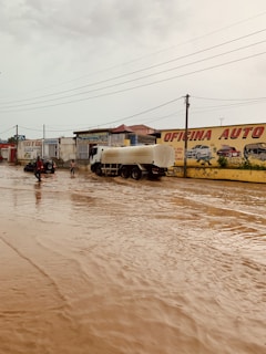 A large water tanker is parked on a flooded street in front of several buildings, one of which has a sign saying 'Oficina Auto'. People are walking through the water, and the sky is overcast.