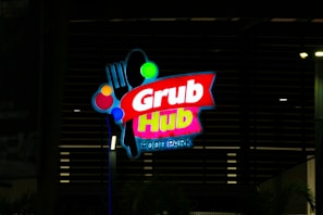 A brightly lit sign for Grub Hub Food Park, featuring the brand name in vibrant colors against a dark background. The sign includes graphic elements such as a fork and circles in various colors.