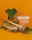A container of honey-flavored cream is placed on a wooden slice, accompanied by a wooden honey dipper, pieces of bread, and a small leaf plant in the background. The scene is set against a bright orange backdrop which complements the packaging of the cream.