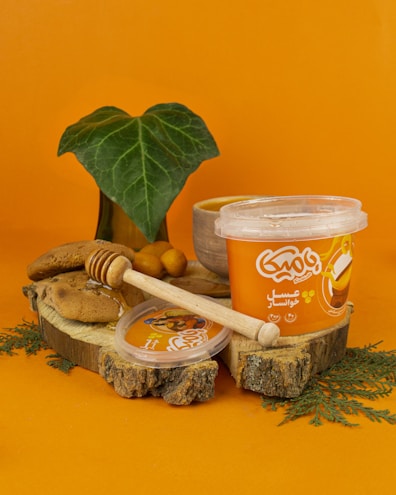 A container of honey-flavored cream is placed on a wooden slice, accompanied by a wooden honey dipper, pieces of bread, and a small leaf plant in the background. The scene is set against a bright orange backdrop which complements the packaging of the cream.