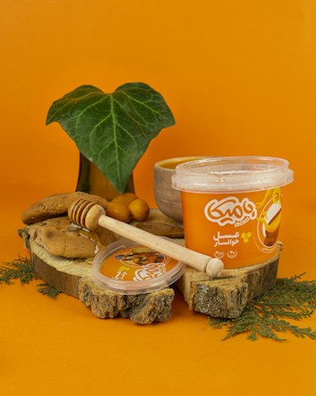 A container of honey-flavored cream is placed on a wooden slice, accompanied by a wooden honey dipper, pieces of bread, and a small leaf plant in the background. The scene is set against a bright orange backdrop which complements the packaging of the cream.