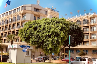 A multi-story hotel with a beige facade and multiple balconies is situated behind a large, lush tree. The building features signage for Golden Tulip, with flags above. A small white kiosk is visible in front with signs in Arabic and English. Several cars are parked along the street, and there is a traffic light nearby.