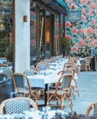 An outdoor dining area features several tables covered with white tablecloths, set with glassware, and surrounded by wicker chairs. The setting is beside a building with a floral patterned exterior wall dominated by large pink flowers and green foliage. A sign displays the name of the establishment.