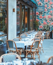 An outdoor dining area features several tables covered with white tablecloths, set with glassware, and surrounded by wicker chairs. The setting is beside a building with a floral patterned exterior wall dominated by large pink flowers and green foliage. A sign displays the name of the establishment.