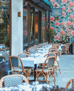 An outdoor dining area features several tables covered with white tablecloths, set with glassware, and surrounded by wicker chairs. The setting is beside a building with a floral patterned exterior wall dominated by large pink flowers and green foliage. A sign displays the name of the establishment.