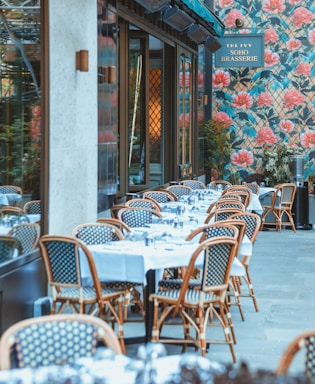 An outdoor dining area features several tables covered with white tablecloths, set with glassware, and surrounded by wicker chairs. The setting is beside a building with a floral patterned exterior wall dominated by large pink flowers and green foliage. A sign displays the name of the establishment.
