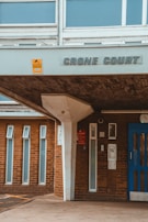 Entrance of a building named 'Crone Court' with a blue door and brick facade. There are several narrow vertical windows and an overhang supported by a white column. A yellow sign is posted near the name signage.