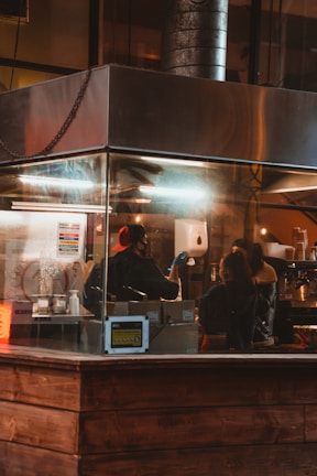 A cozy cafe setting with warm lighting dominates the scene. Two individuals are inside a small, enclosed area with glass walls, working behind a counter. There are various coffee-making equipment and supplies visible. The ambiance suggests an inviting and bustling atmosphere typical of a coffee shop.