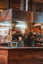 Inside a kitchen or restaurant setting, a person in a red hat is standing behind a counter, interacting with another person. The area is enclosed in metal and glass structures, with equipment such as a dispenser and various cooking utensils visible. Warm lighting illuminates the scene, creating a cozy atmosphere.