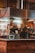 Inside a kitchen or restaurant setting, a person in a red hat is standing behind a counter, interacting with another person. The area is enclosed in metal and glass structures, with equipment such as a dispenser and various cooking utensils visible. Warm lighting illuminates the scene, creating a cozy atmosphere.