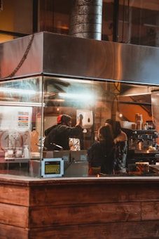 Inside a kitchen or restaurant setting, a person in a red hat is standing behind a counter, interacting with another person. The area is enclosed in metal and glass structures, with equipment such as a dispenser and various cooking utensils visible. Warm lighting illuminates the scene, creating a cozy atmosphere.