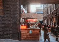 A cozy alleyway features a small food stall with a bright red neon sign reading 'BBQ'. The stall is made of wood and has an industrial-style vent. People are gathered around the stall and seated at outdoor tables, enjoying food and conversation. String lights are hung across the alley, and the surrounding walls are made of dark and brown brick.