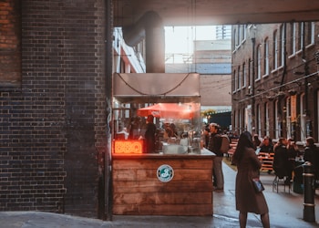 A cozy alleyway features a small food stall with a bright red neon sign reading 'BBQ'. The stall is made of wood and has an industrial-style vent. People are gathered around the stall and seated at outdoor tables, enjoying food and conversation. String lights are hung across the alley, and the surrounding walls are made of dark and brown brick.