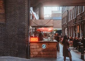 A cozy alleyway features a small food stall with a bright red neon sign reading 'BBQ'. The stall is made of wood and has an industrial-style vent. People are gathered around the stall and seated at outdoor tables, enjoying food and conversation. String lights are hung across the alley, and the surrounding walls are made of dark and brown brick.