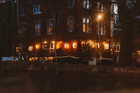 A street scene at night features a brick building adorned with warm, illuminated lanterns. String lights add a cozy ambiance, and a large tree partially obscures part of the view. The windows glow softly, suggesting activity inside.