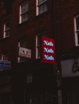 A dimly lit street scene features a series of signs attached to brick buildings. A prominent red sign with the word 'Nails' repeated three times in light letters stands out among other signs for a tailor and dry cleaner. The buildings give a sense of an urban environment.