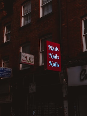 A dimly lit street scene features a series of signs attached to brick buildings. A prominent red sign with the word 'Nails' repeated three times in light letters stands out among other signs for a tailor and dry cleaner. The buildings give a sense of an urban environment.