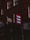 A dimly lit street scene features a series of signs attached to brick buildings. A prominent red sign with the word 'Nails' repeated three times in light letters stands out among other signs for a tailor and dry cleaner. The buildings give a sense of an urban environment.