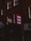 A dimly lit street scene features a series of signs attached to brick buildings. A prominent red sign with the word 'Nails' repeated three times in light letters stands out among other signs for a tailor and dry cleaner. The buildings give a sense of an urban environment.