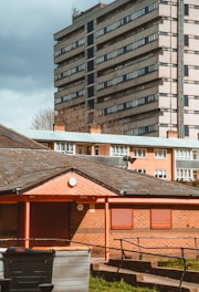 A residential area with a foreground of a small brick building featuring red shutters and a sloped roof. In the mid-ground, there are several large waste bins, and a taller apartment building with numerous windows is in the background. The sky is overcast, giving the scene a muted lighting.
