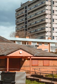 A residential area with a foreground of a small brick building featuring red shutters and a sloped roof. In the mid-ground, there are several large waste bins, and a taller apartment building with numerous windows is in the background. The sky is overcast, giving the scene a muted lighting.