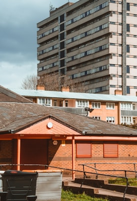A residential area with a foreground of a small brick building featuring red shutters and a sloped roof. In the mid-ground, there are several large waste bins, and a taller apartment building with numerous windows is in the background. The sky is overcast, giving the scene a muted lighting.