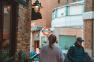 A street scene with a focus on two people walking. A woman with a ponytail is wearing a light sweater, and a man wearing a cap and a padded jacket is carrying a backpack. They are near a brick building with a lamp on its exterior, and a London Underground sign is visible in the background.