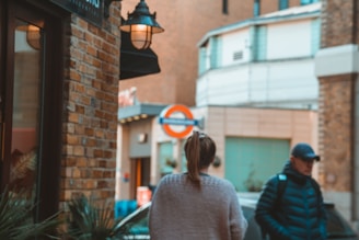 A street scene with a focus on two people walking. A woman with a ponytail is wearing a light sweater, and a man wearing a cap and a padded jacket is carrying a backpack. They are near a brick building with a lamp on its exterior, and a London Underground sign is visible in the background.