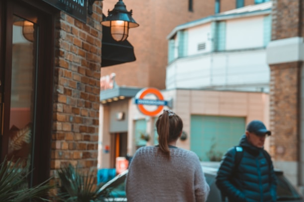 A street scene with a focus on two people walking. A woman with a ponytail is wearing a light sweater, and a man wearing a cap and a padded jacket is carrying a backpack. They are near a brick building with a lamp on its exterior, and a London Underground sign is visible in the background.