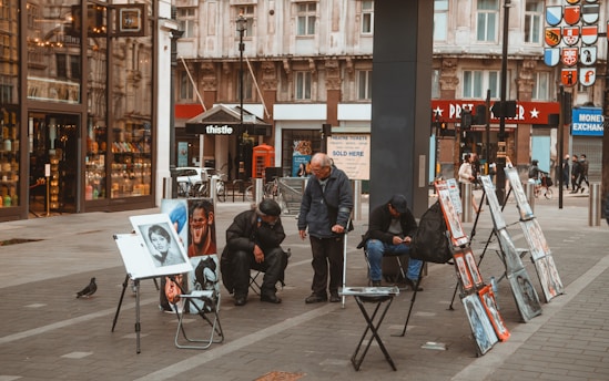 Street scene with artists displaying portraits and caricatures on easels. Three people are gathered around the artwork and a passerby walks by in the background. The backdrop features urban buildings, a red phone booth, and storefronts. A pigeon is seen on the pavement.