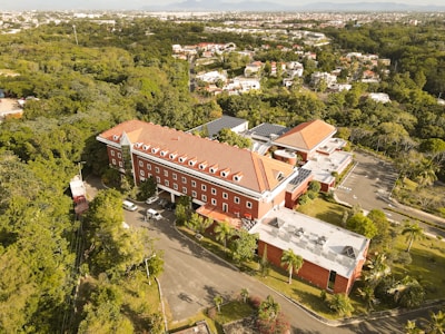 An aerial view of a large, red-roofed building surrounded by lush green trees. The building is set in a spacious area with a parking lot and access roads. Residential areas with smaller houses can be seen in the background, and there are distant mountains on the horizon.