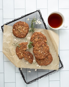 A set of oat cookies with visible grains, arranged on parchment paper, atop a dark wire tray. Fresh rosemary sprigs are scattered among the cookies, and a small amount of rolled oats are sprinkled around. Next to the tray is a white cup filled with tea, situated on a tiled surface.