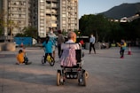 A variety of people are gathered in an outdoor urban area near a high-rise building. In the foreground, an elderly person is seated in a wheelchair, facing away. Several children are playing nearby, one on a bicycle and another on a scooter. There are adults in the background, some of whom are wearing masks.