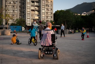 A variety of people are gathered in an outdoor urban area near a high-rise building. In the foreground, an elderly person is seated in a wheelchair, facing away. Several children are playing nearby, one on a bicycle and another on a scooter. There are adults in the background, some of whom are wearing masks.