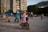 A variety of people are gathered in an outdoor urban area near a high-rise building. In the foreground, an elderly person is seated in a wheelchair, facing away. Several children are playing nearby, one on a bicycle and another on a scooter. There are adults in the background, some of whom are wearing masks.