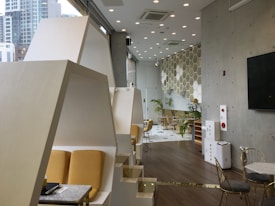 A modern interior space with geometric and minimalist design elements. The room features hexagonal wall decorations, large windows revealing an urban skyline, and yellow cushioned seating areas. There are potted plants adding a touch of greenery and the flooring is a dark wood. Lighting is ample, with numerous ceiling lights contributing to a bright atmosphere.