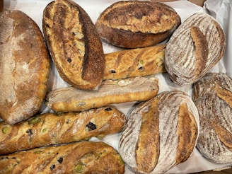 A selection of various artisanal breads placed on a surface. The loaves include different shapes and textures, such as baguettes and round boules, featuring rustic crusts and visible ingredients like olives.